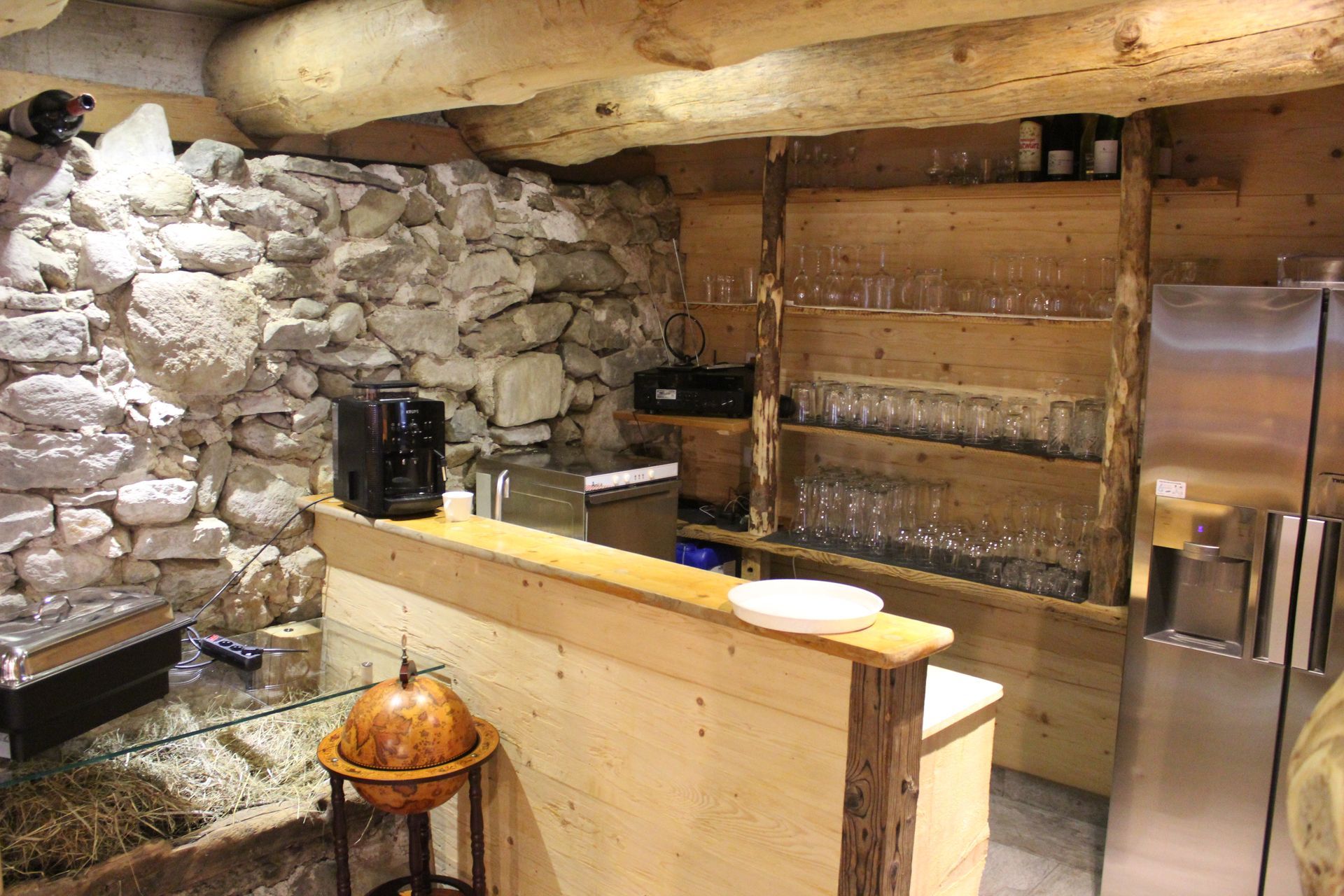 Eine hölzerne Bartheke mit Kaffeemaschine, vor einer Steinwand und Regalen voller Gläser in einem rustikalen Raum.