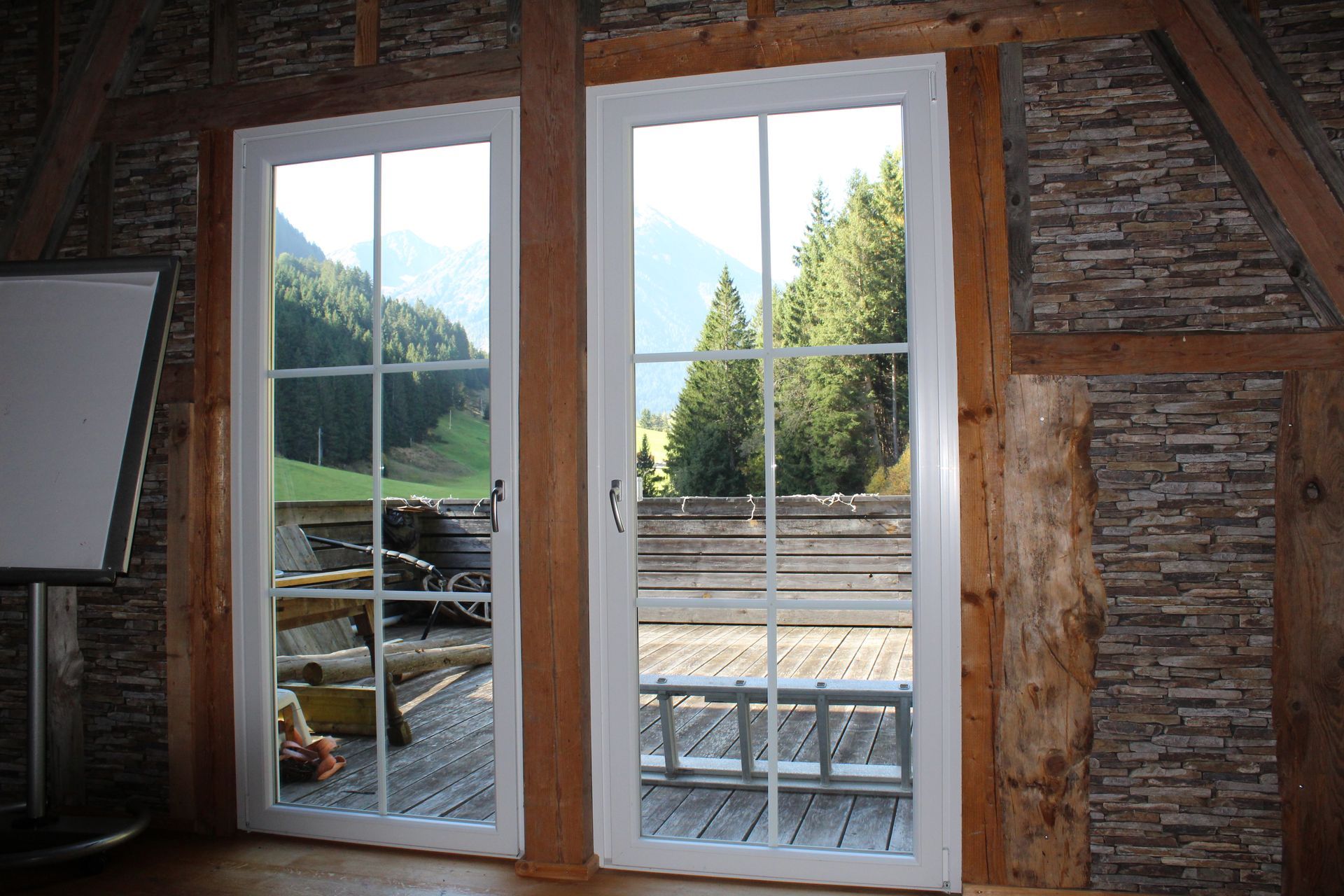 Zwei weißgerahmte Glastüren in einer rustikalen Holzwand bieten Blick auf einen Holzbalkon, Wald und Berge.