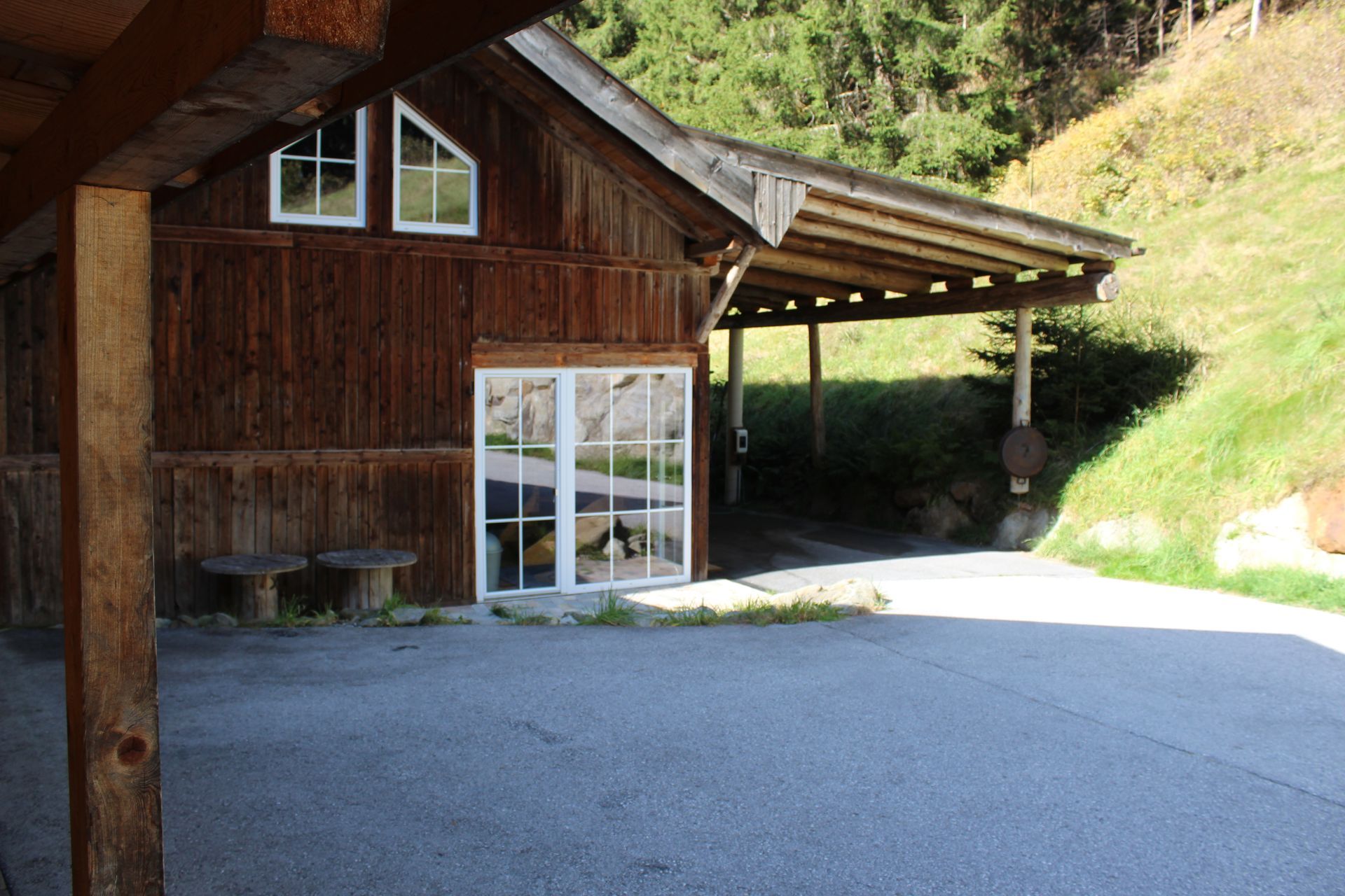 Ein rustikales Holzgebäude mit seitlichem Carport und weiß gerahmten Glastüren, gelegen an einem grasbewachsenen Hang.