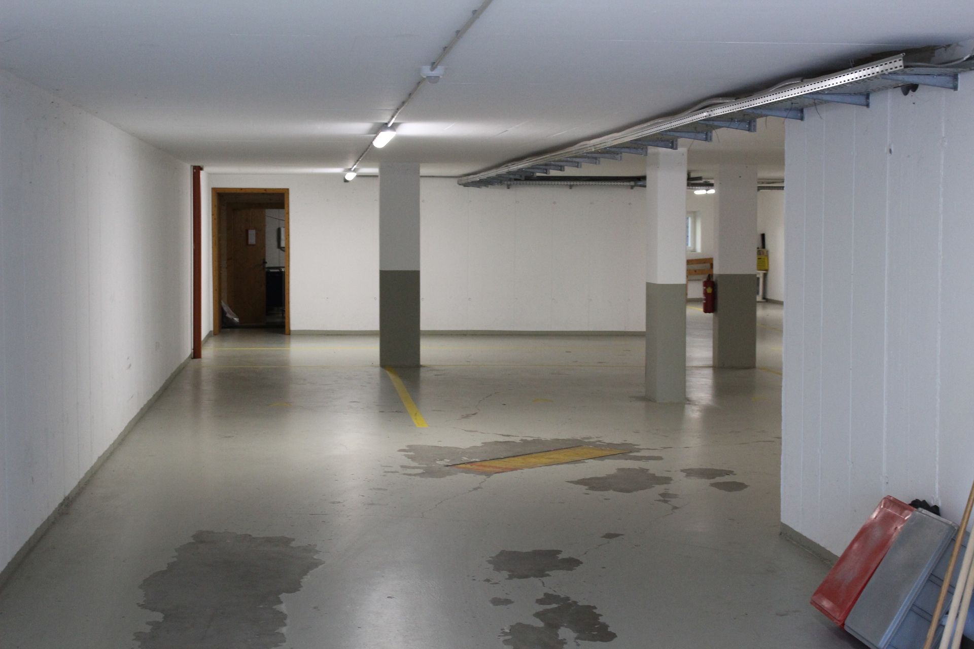 Eine leere Tiefgarage mit Betonboden, weißen Wänden, Stützpfeilern und Deckenbeleuchtung aus Leuchtstoffröhren.