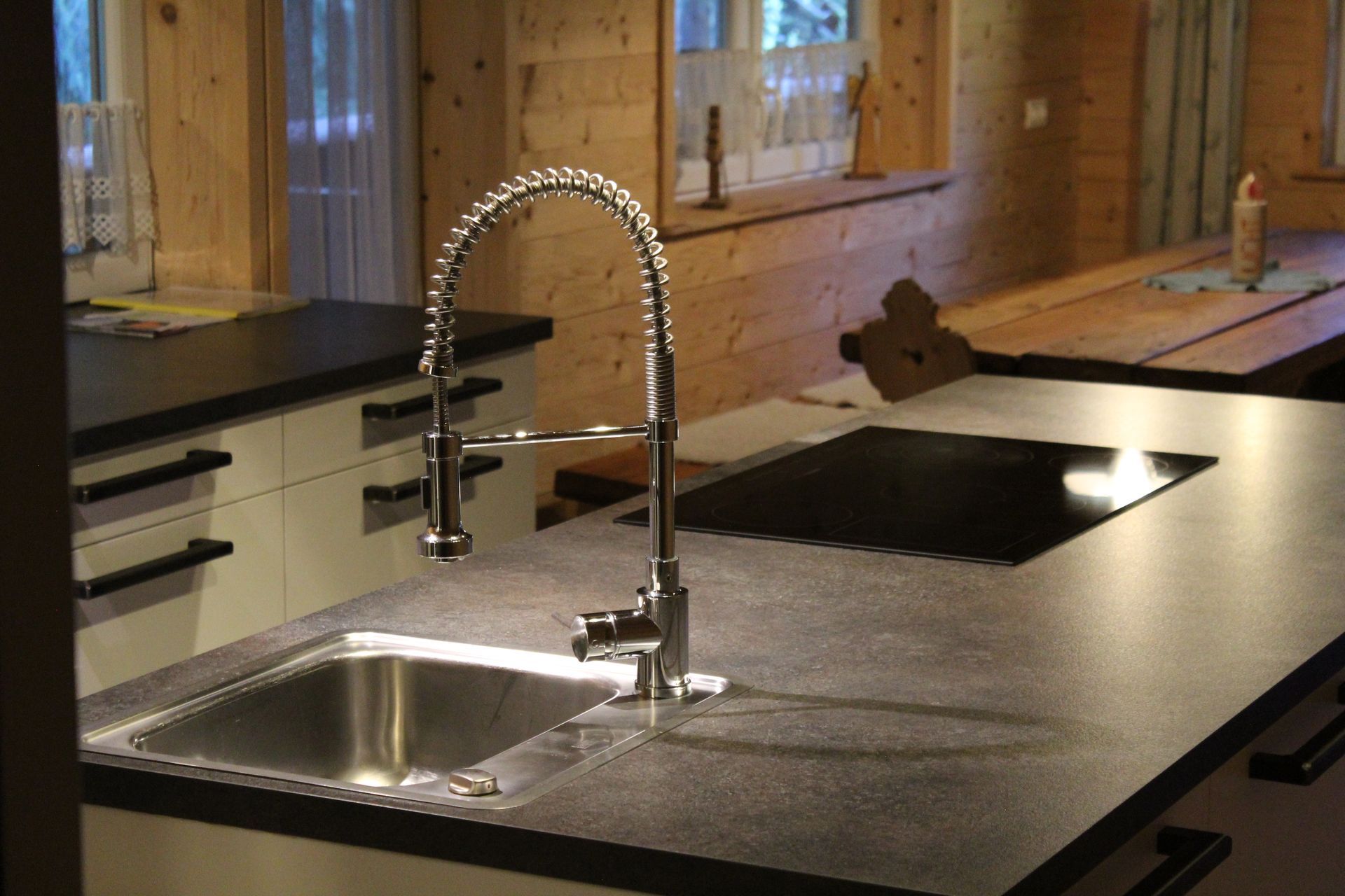 Eine moderne Kücheninsel mit dunkler Arbeitsplatte, Edelstahlspüle, Schwanenhalsarmatur und schwarzem Einbaukochfeld.