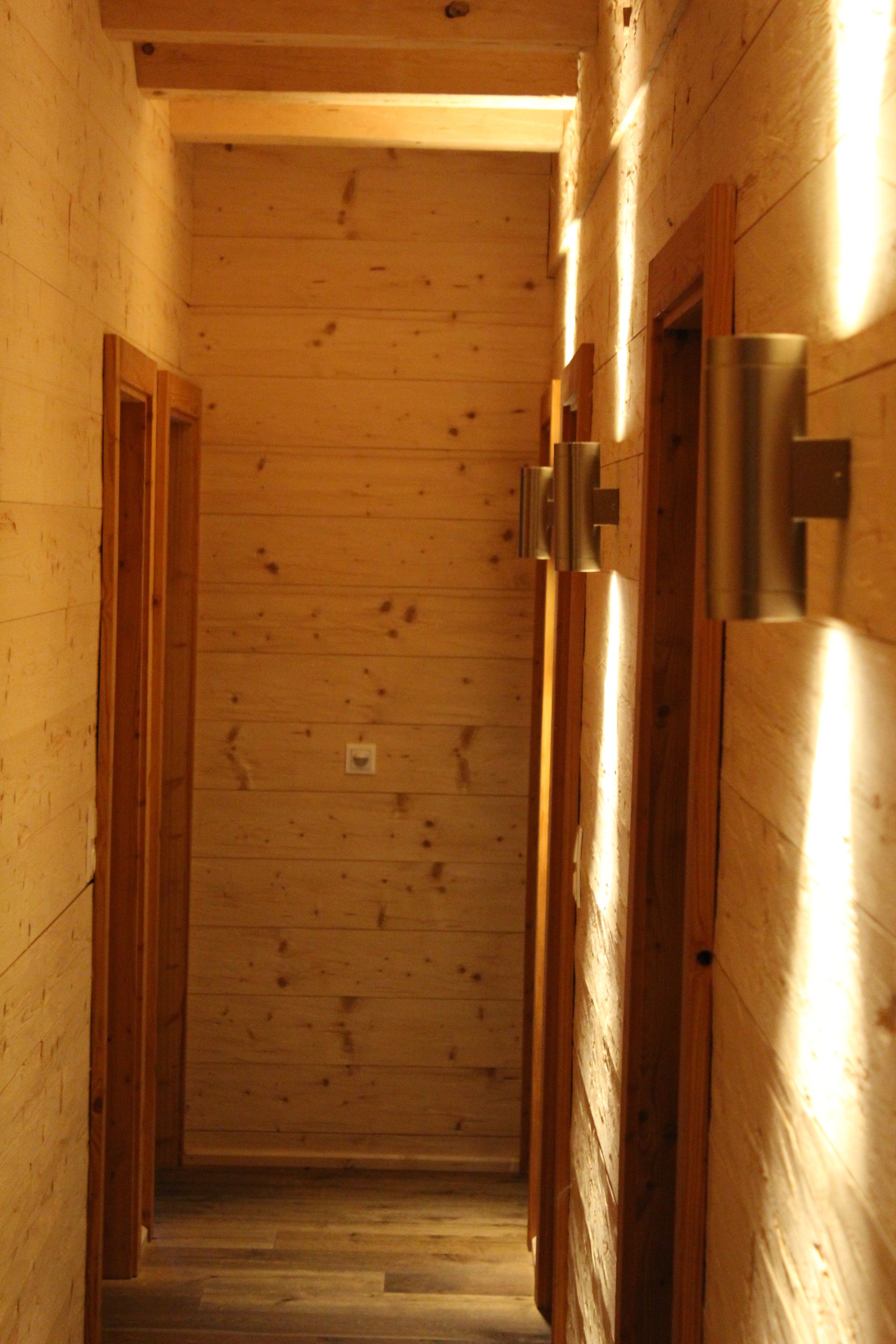 Ein schmaler Flur mit hellen, holzgetäfelten Wänden und Fußboden, der auf der rechten Seite von Wandleuchten beleuchtet wird.