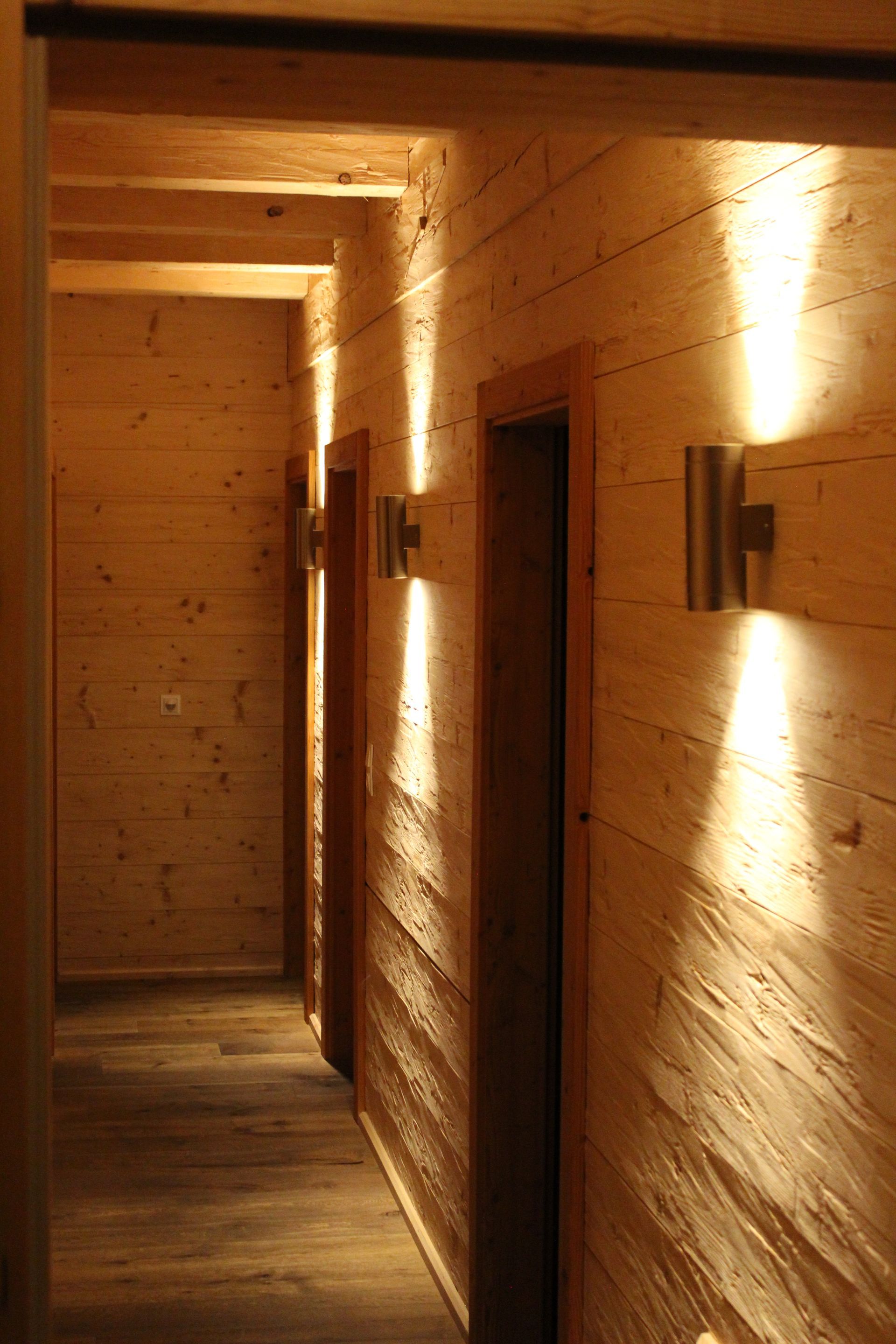 Ein Flur mit warmen Holzwänden und -böden, der von modernen Wandleuchten erhellt wird, die vertikale Lichtstrahlen werfen.