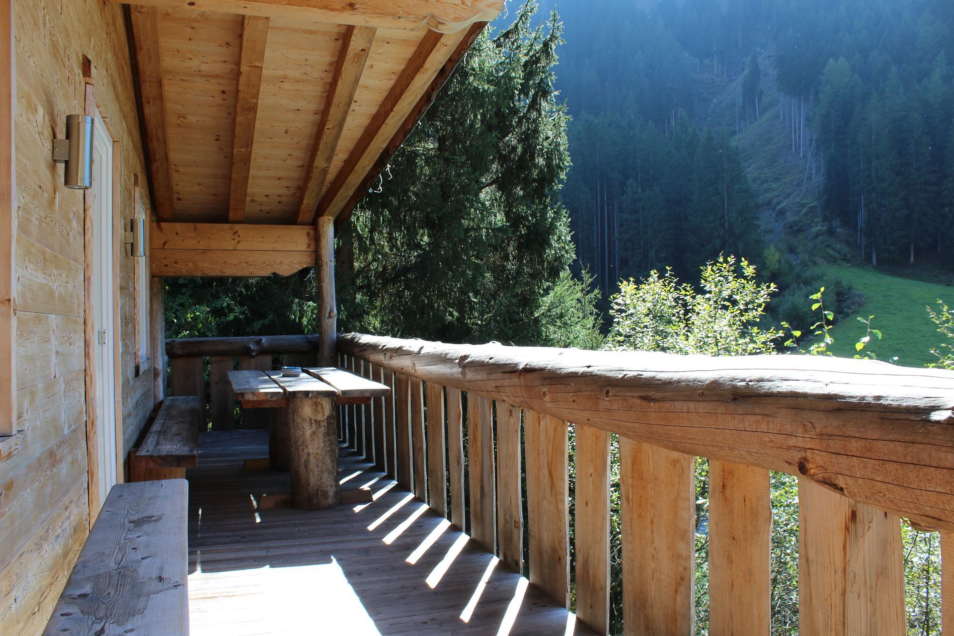 Ein rustikaler Holzbalkon mit Tisch und Bank bietet an einem sonnigen Tag einen herrlichen Blick auf ein grünes Waldtal.