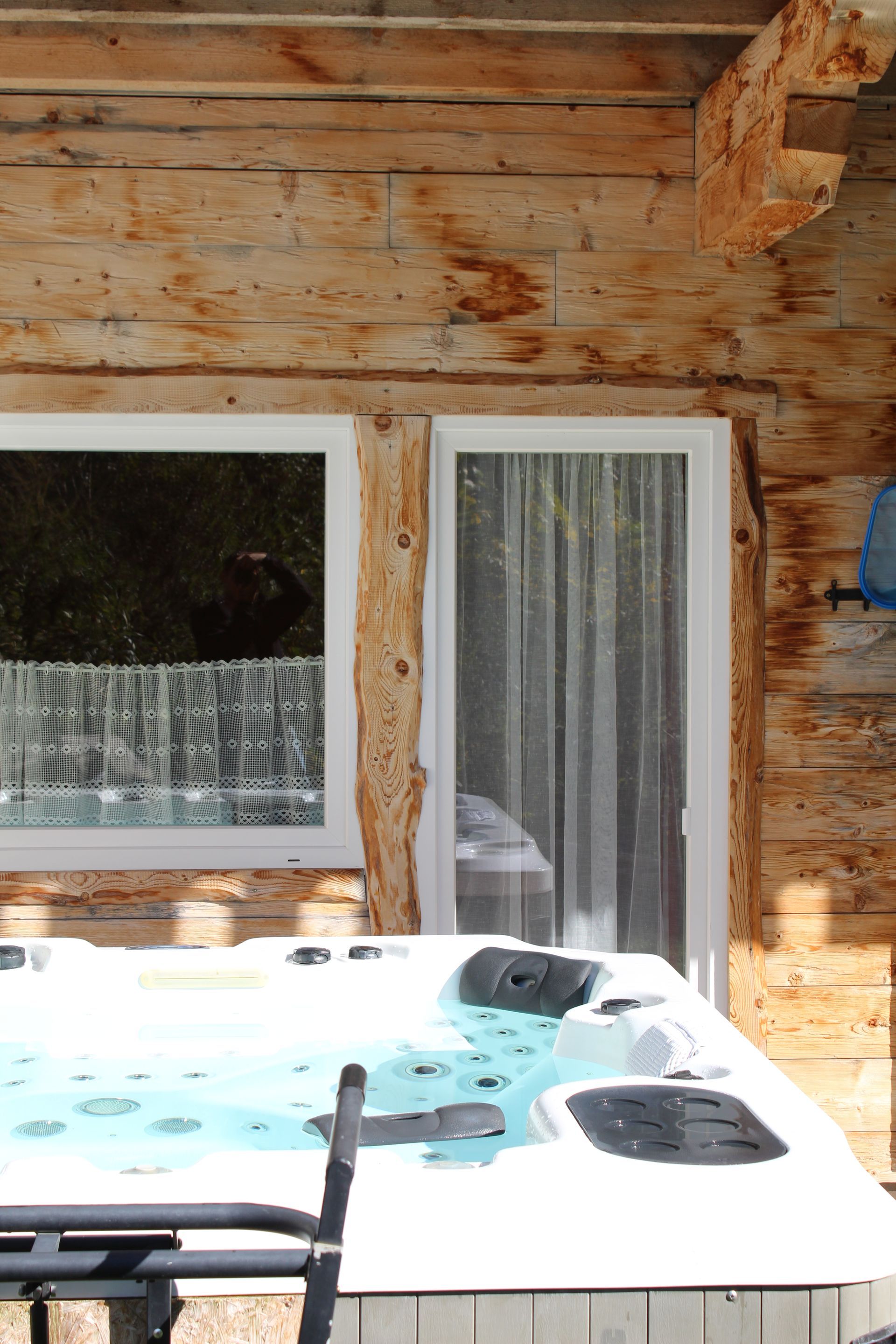 Auf einer Terrasse neben einer Blockhauswand mit einem weiß gerahmten Fenster und einer Glastür steht ein Whirlpool.