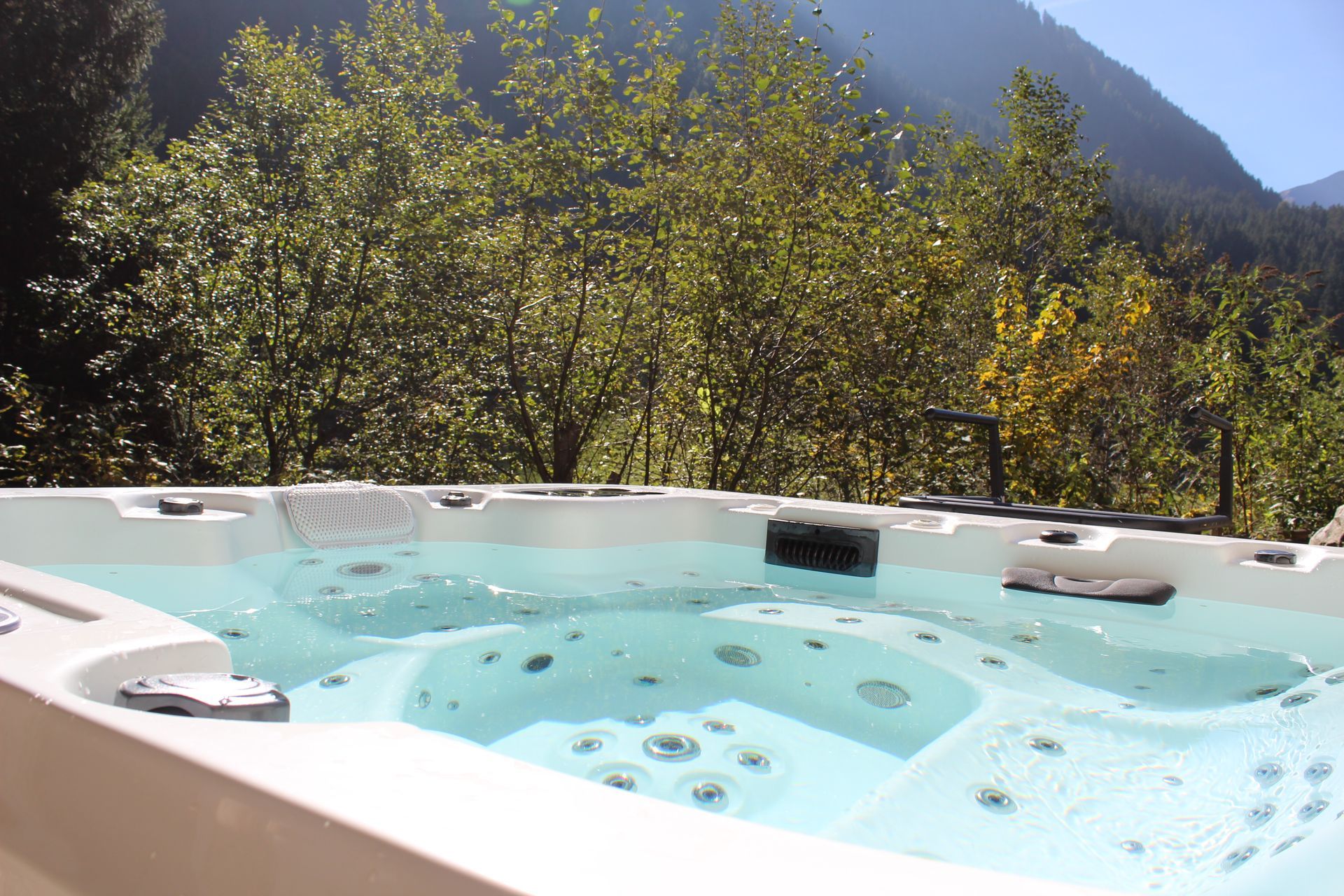 Ein weißer Whirlpool, gefüllt mit klarem Wasser, im Freien gelegen vor dem Hintergrund grüner Bäume und eines Berges.