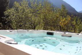 Ein weißer Whirlpool, gefüllt mit klarem Wasser, im Freien gelegen vor dem Hintergrund grüner Bäume und eines Berges.