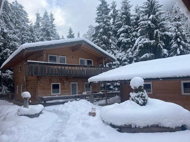 Eine rustikale Holzhütte und ein angrenzendes Gebäude, umgeben von schneebedeckten Kiefern an einem Wintertag.