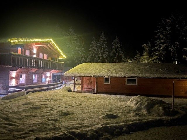 Nachts steht neben einem kleineren, schneebedeckten Gebäude eine zweistöckige Holzhütte