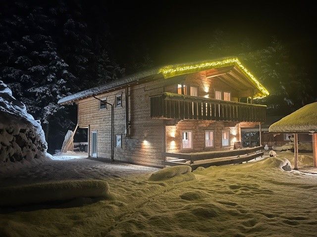 Eine zweistöckige Holzhütte in der Nacht, beleuchtet von gelben Dach- und Wandlampen, inmitten einer verschneiten Landschaft.