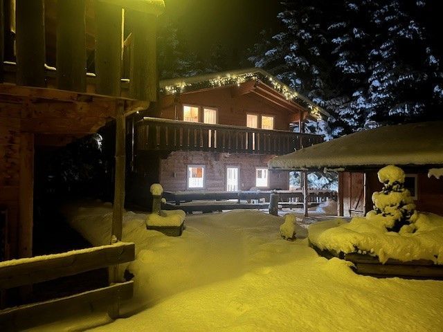 Eine hölzerne Hütte, nachts von warmen Lichtern erhellt, steht in einem schneebedeckten Hof, umgeben von dunklen Bäumen.