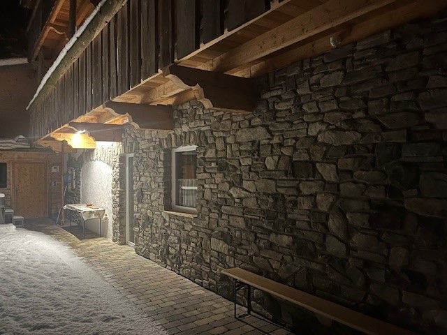 Nachtansicht einer steinernen Berghütte mit Holzbalkon, die von warmen Lichtern über einem verschneiten Pfad beleuchtet wird.
