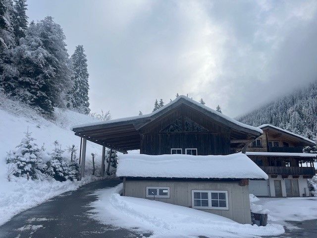Eine schneebedeckte Hütte mit dunkler Holzfassade liegt in einer verschneiten Berglandschaft unter einem bewölkten Himmel.
