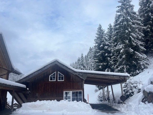Unter einem schneebedeckten Dach steht ein Holzchalet; im Hintergrund sind Kiefern und im Vordergrund tiefer Schnee zu sehen.