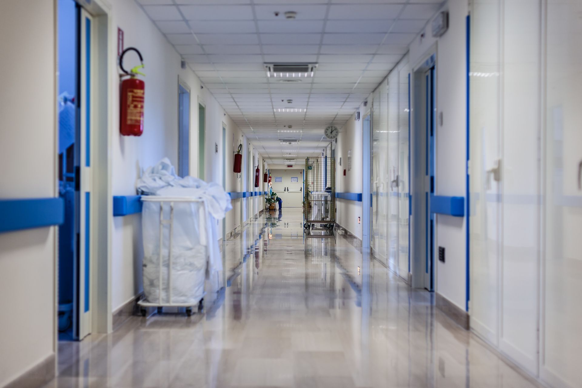 A long, empty hospital corridor with white walls, blue trim, a red fire extinguisher, and a laundry cart on the left.