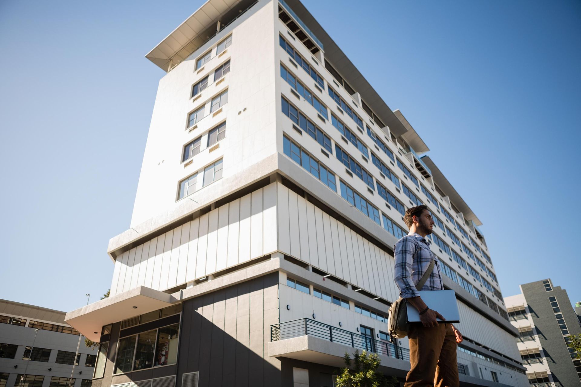 A man is standing in front of a large building holding a laptop.