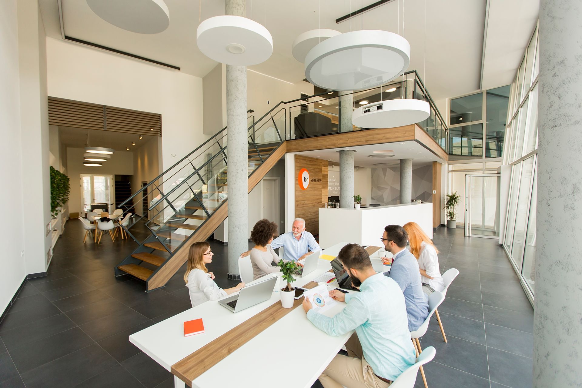Office meeting at a long white table with people, a laptop, and documents. Staircase and upper level in the background.