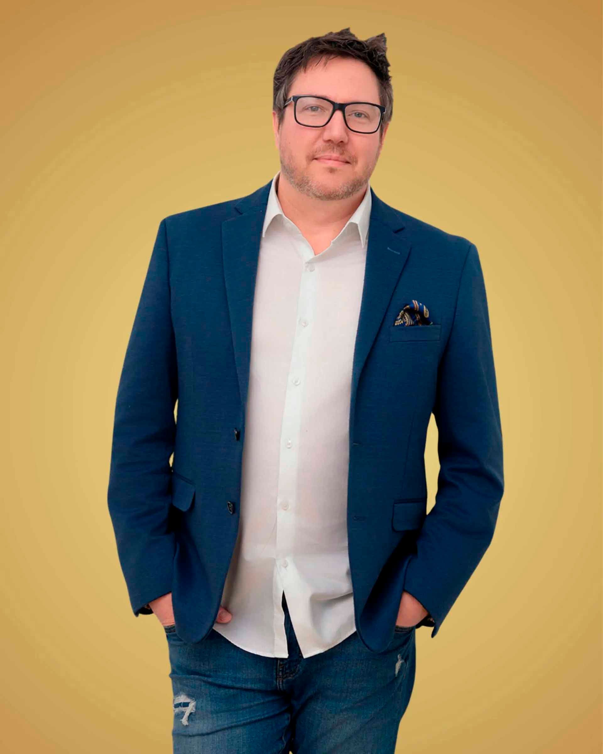 Homme en blazer bleu, chemise blanche et jean, mains dans les poches, souriant. Fond jaune.