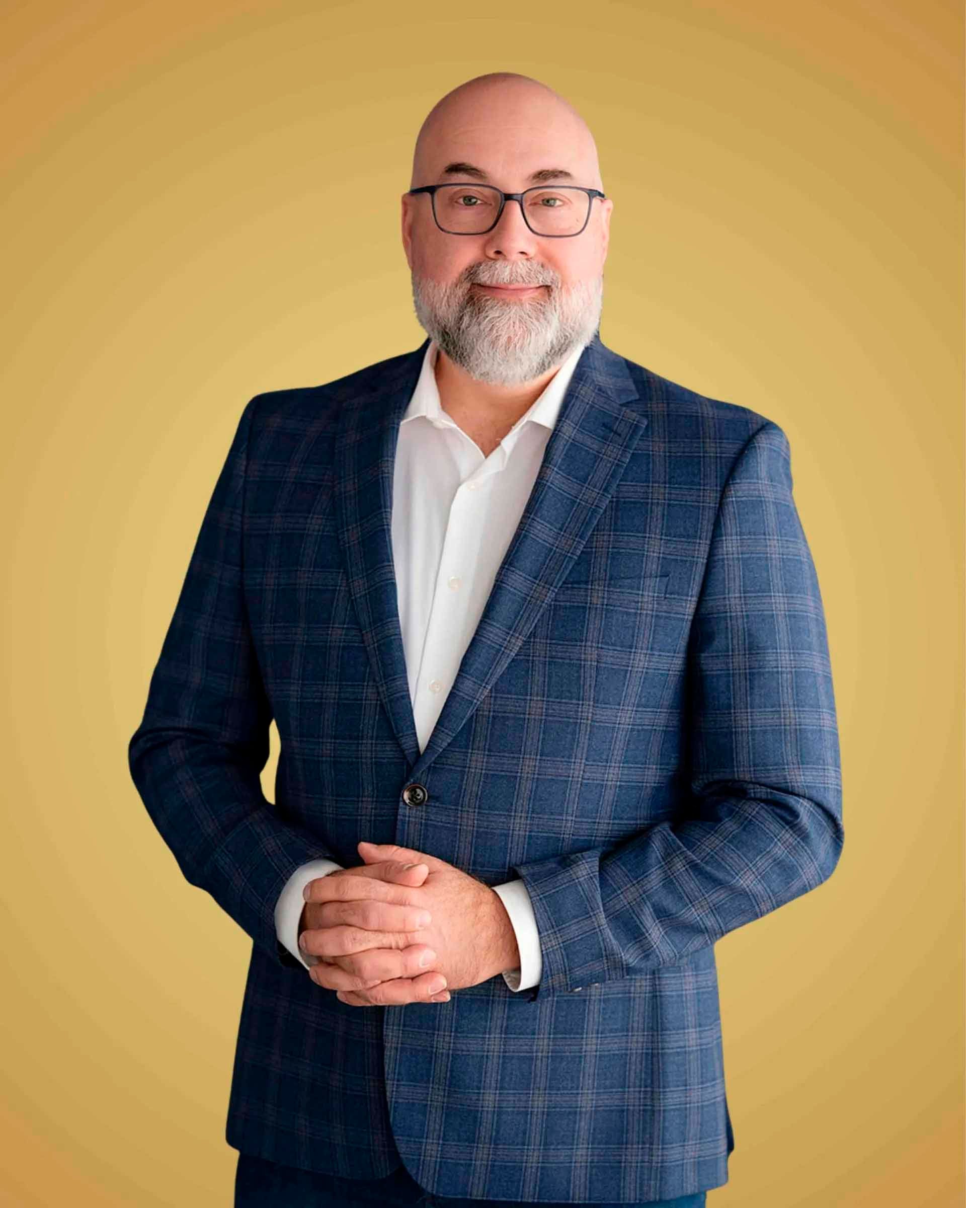 Un homme portant des lunettes et une barbe, vêtu d'un blazer bleu, d'une chemise blanche et d'un jean, se tient devant un fond jaune, les mains jointes.