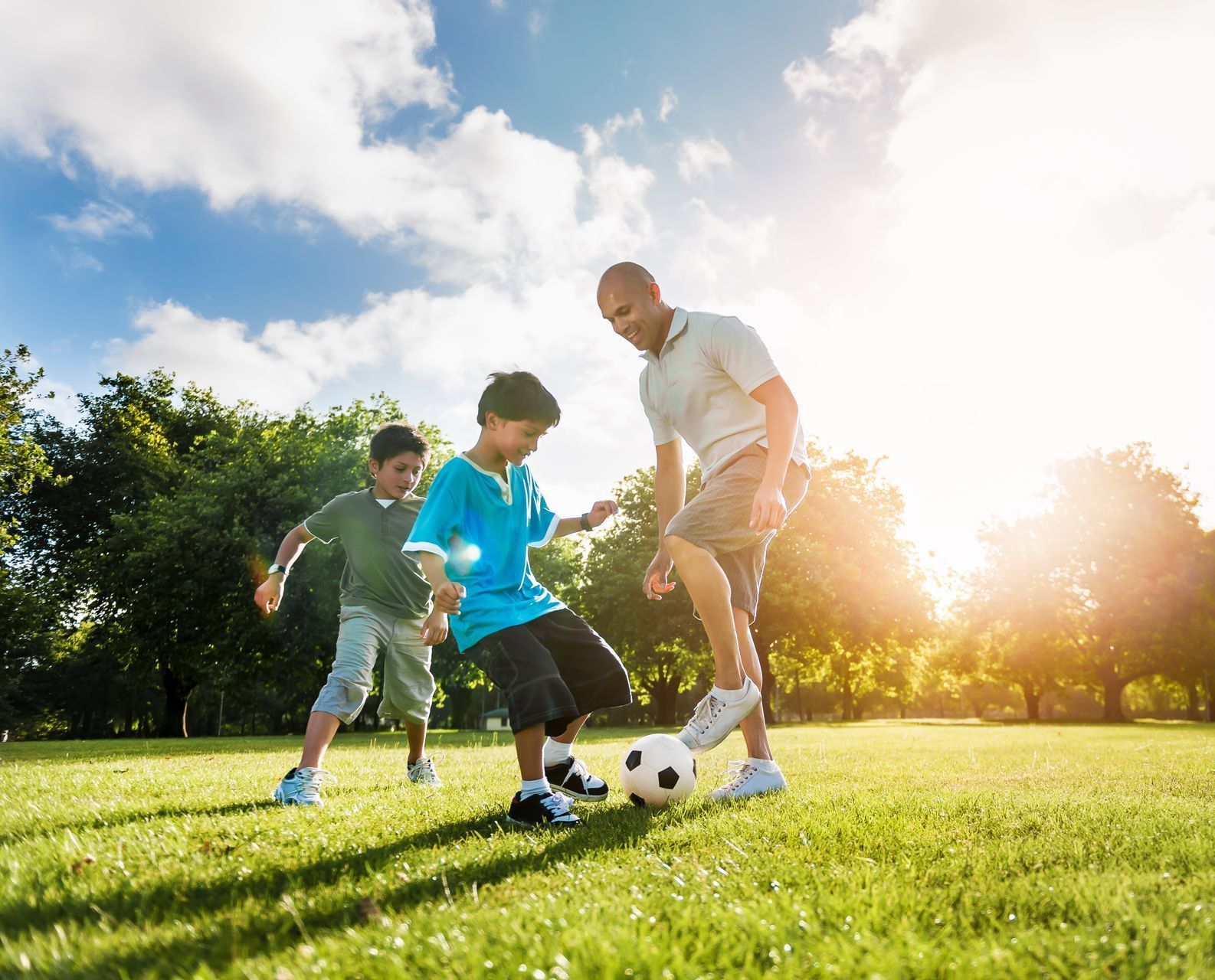 Father and two children playing soccer on a sunny, grassy field.
