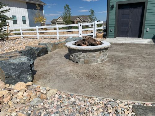Stone fire pit with firewood in a backyard patio area, near a fence and house.
