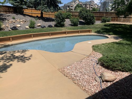 Backyard with a light blue pool, tan patio, and landscaping with grass, rocks, and wooden fencing.