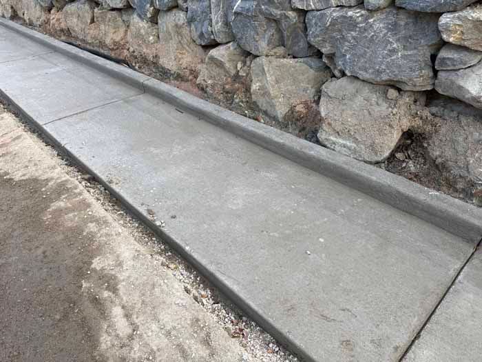 Concrete gutter next to a stone wall, on a paved surface.