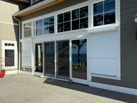 A modern house exterior featuring floor-to-ceiling glass doors and windows, some partially covered by white shutters.