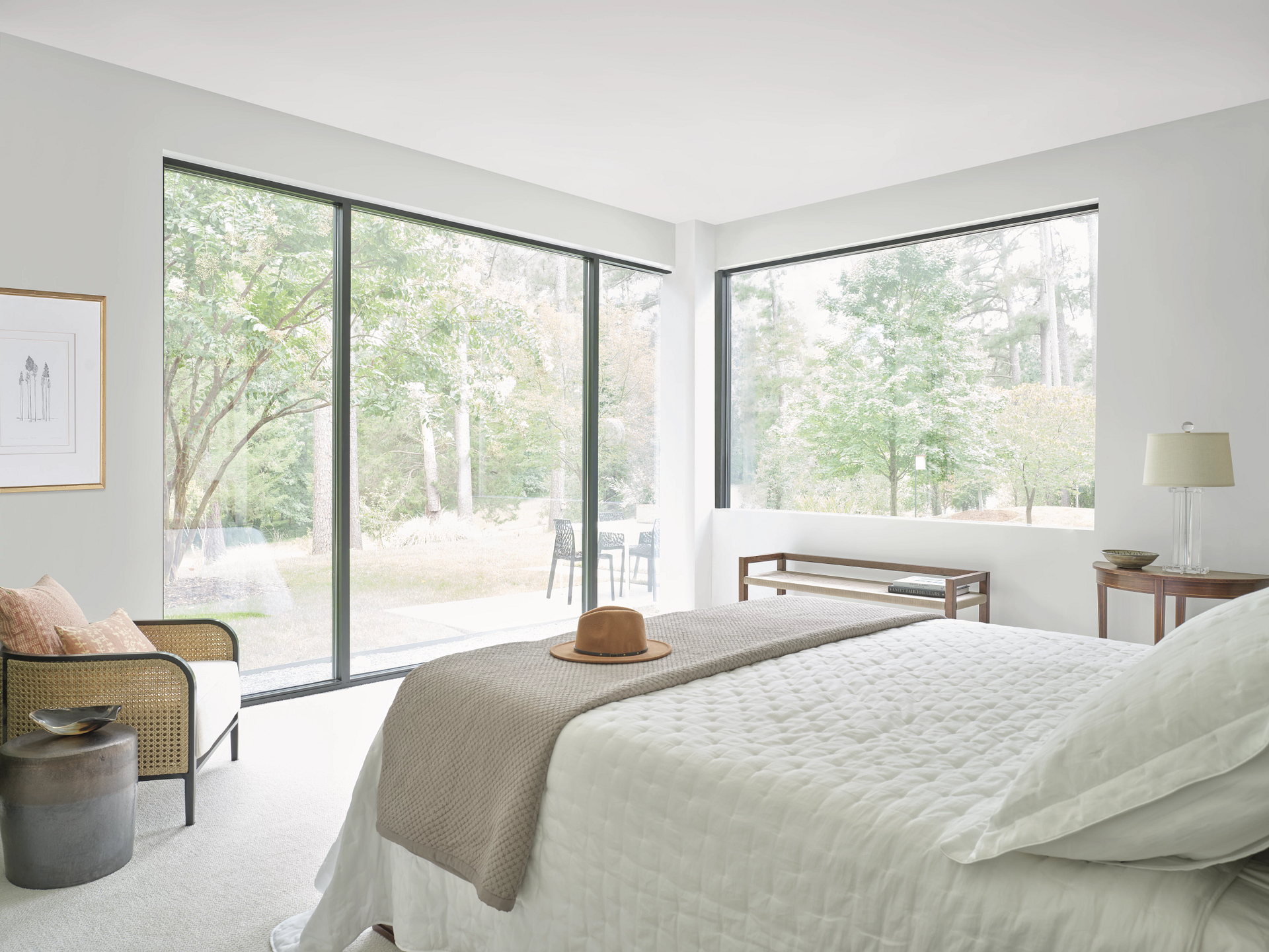 A bright, modern bedroom featuring a bed with white bedding, a straw hat, a side chair, and large windows with forest views.