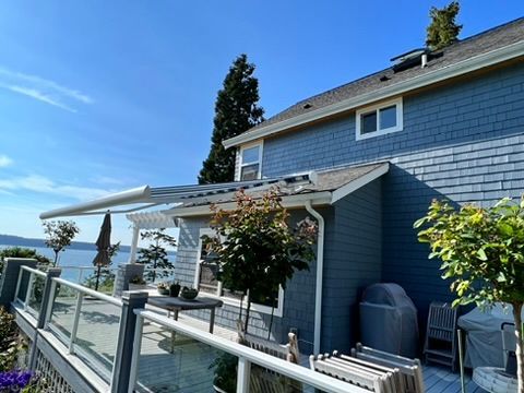 A blue, shingled two-story house with a white pergola over a deck overlooking the ocean on a clear, sunny day.