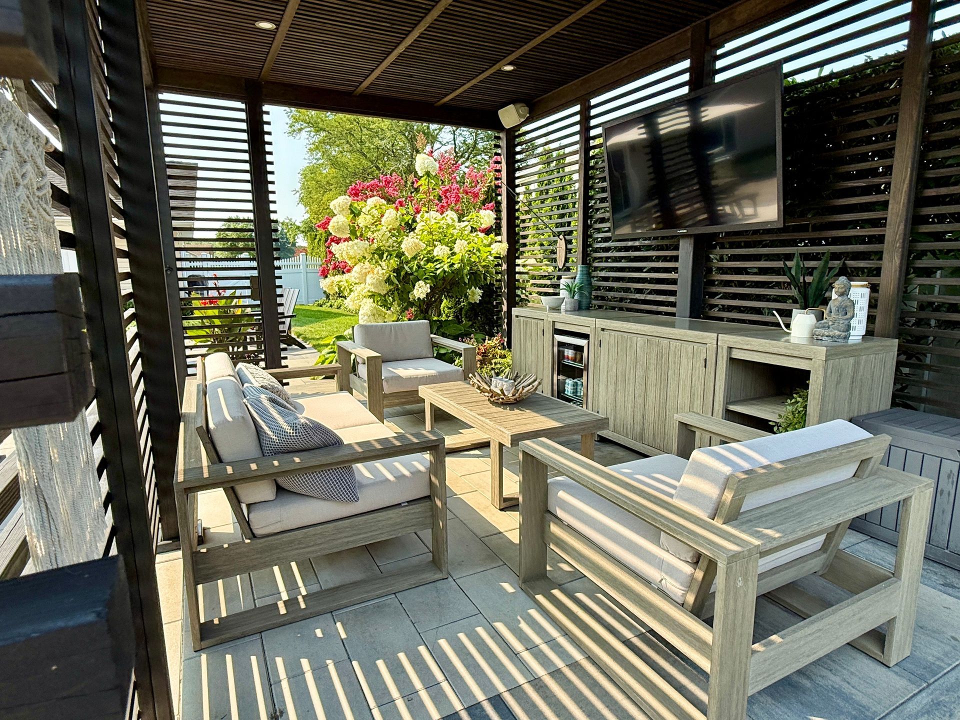 Outdoor patio lounge with light wood furniture, a sofa, coffee table, TV, and mini-fridge under a slatted wood pergola.
