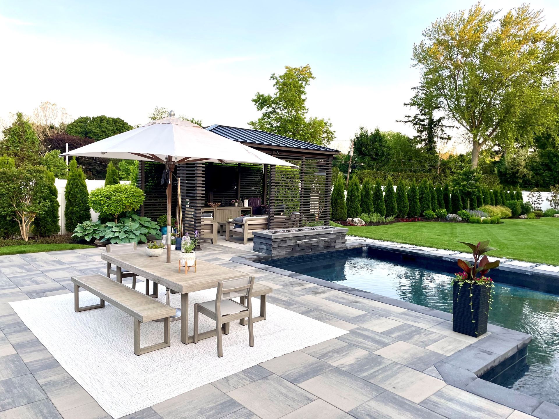An outdoor patio with a dining table and white umbrella, a swimming pool, and a shaded pergola structure on a sunny day.