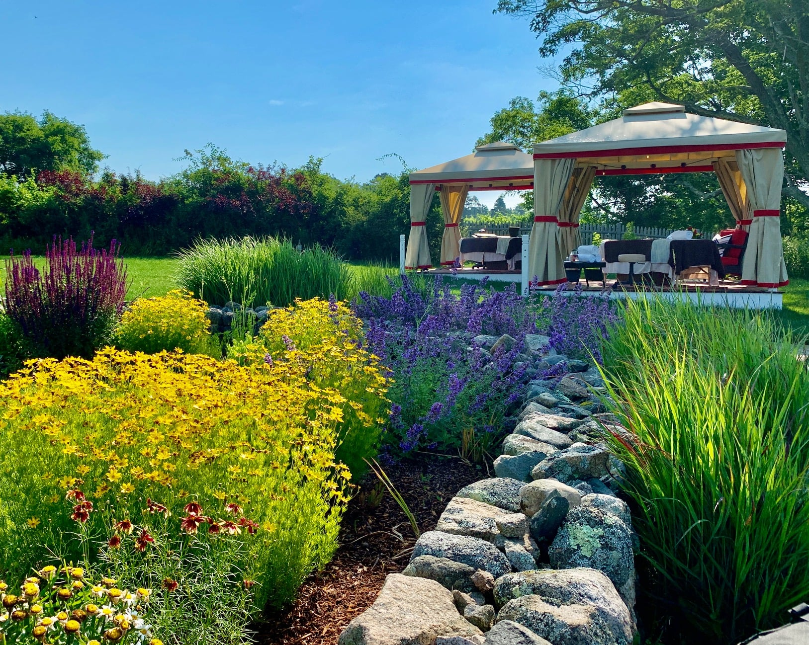 Vibrant garden with yellow and purple flowers, a stone path, and two beige gazebos under a clear blue sky.