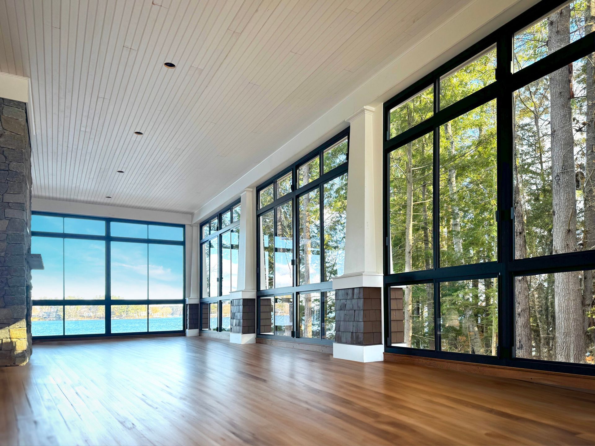 Empty room with large windows overlooking a forest and water. Wooden floor, white ceiling, stone accents.