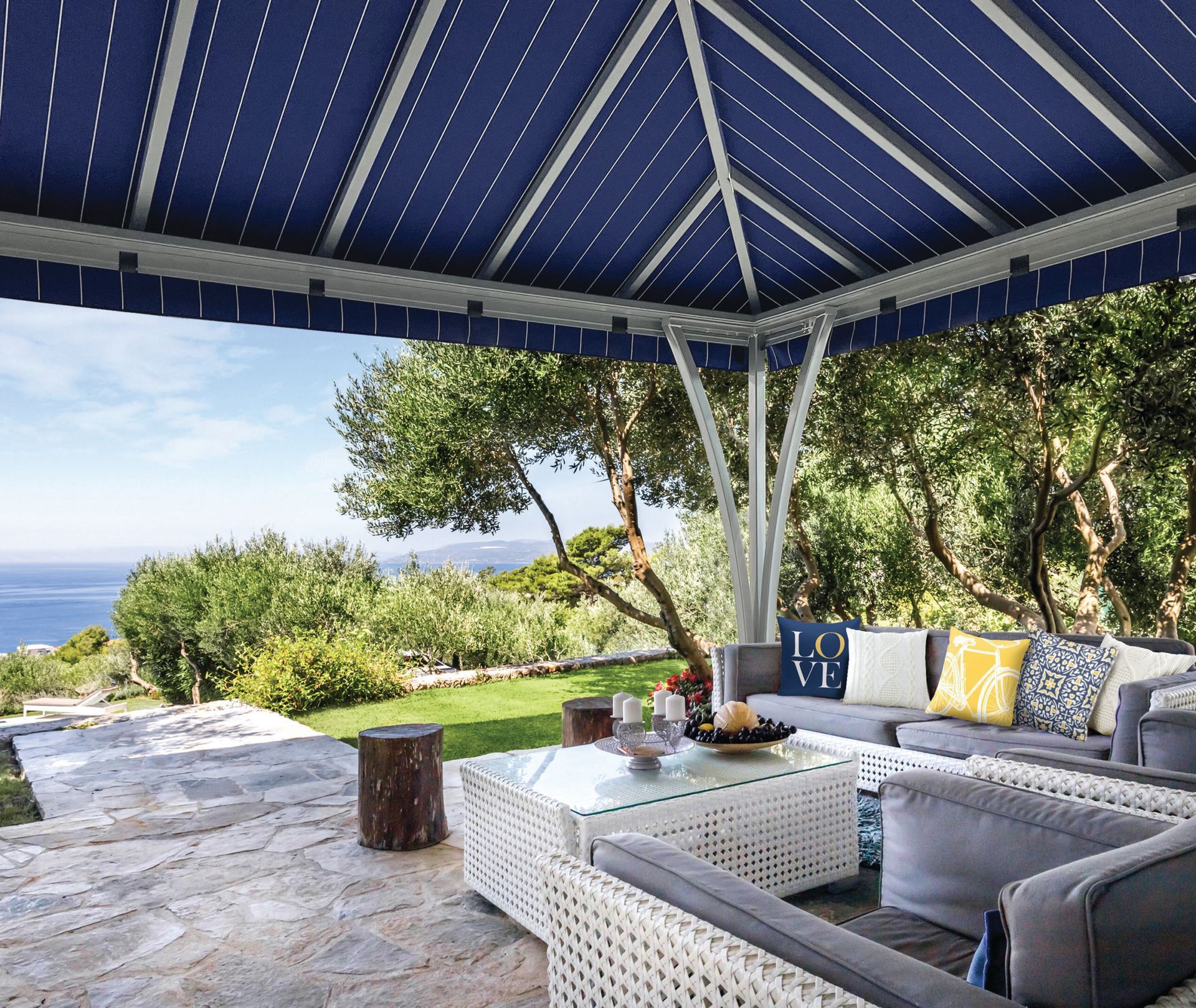 Patio with a blue striped gazebo covering a gray sofa and white coffee table, overlooking a lush garden and the sea.
