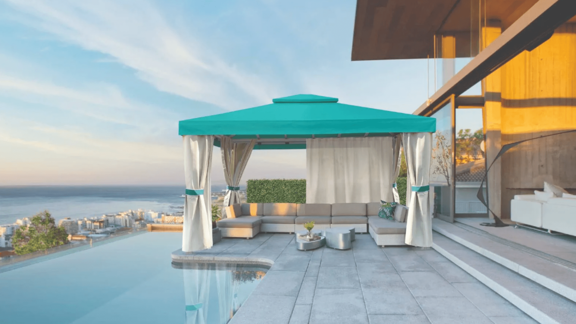 A teal gazebo with white curtains sits on a patio by an infinity pool overlooking a coastal city at sunset.