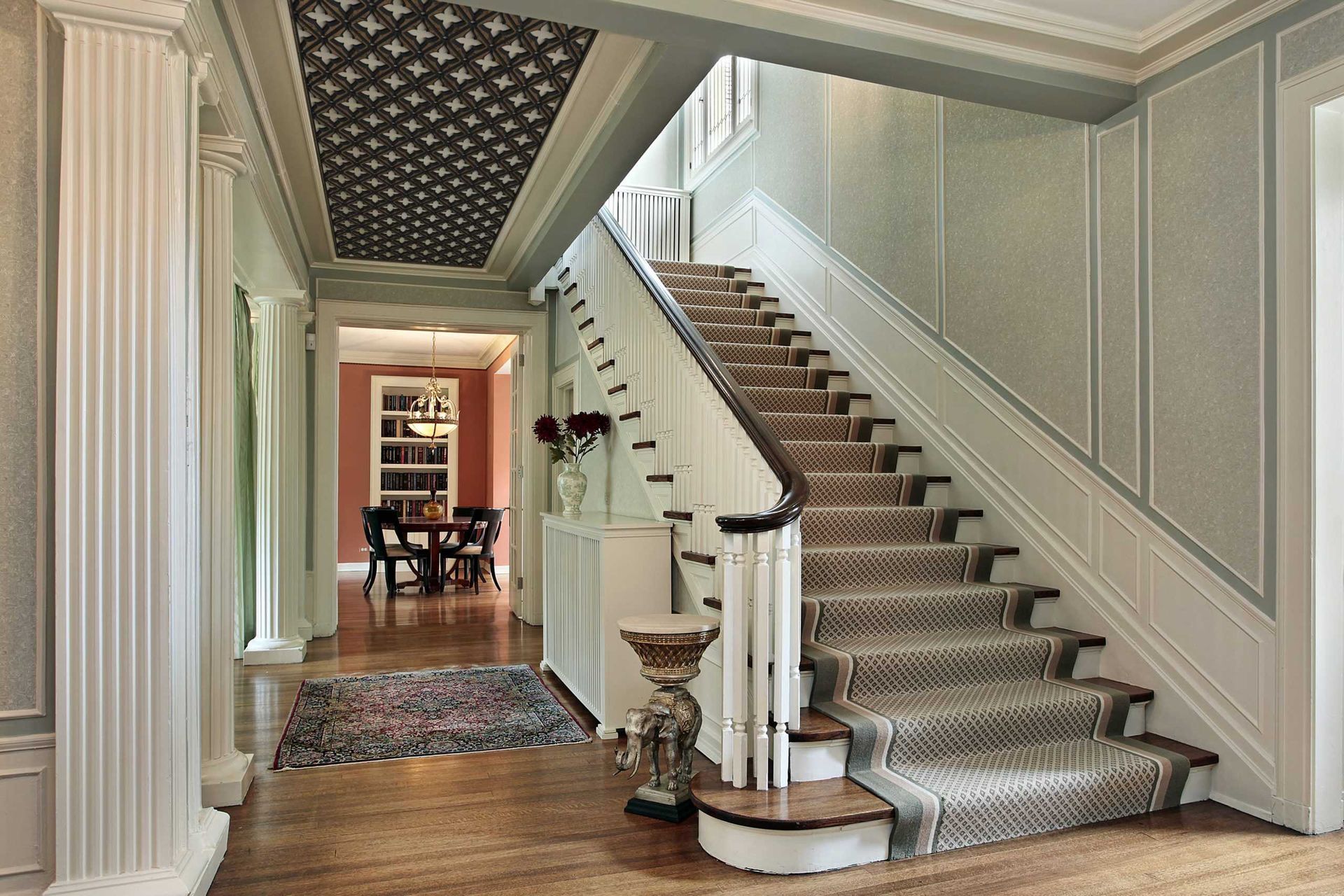 A foyer featuring a carpeted staircase with a white railing, leading to a dining room with a patterned ceiling.