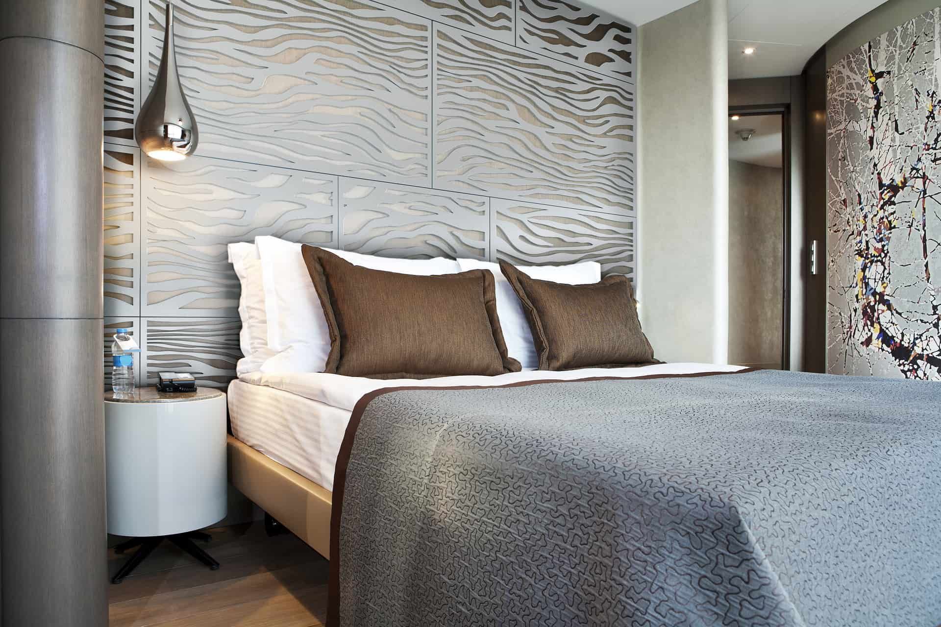 A modern hotel bedroom with a queen bed, grey patterned headboard wall, pendant light, and a small white side table.