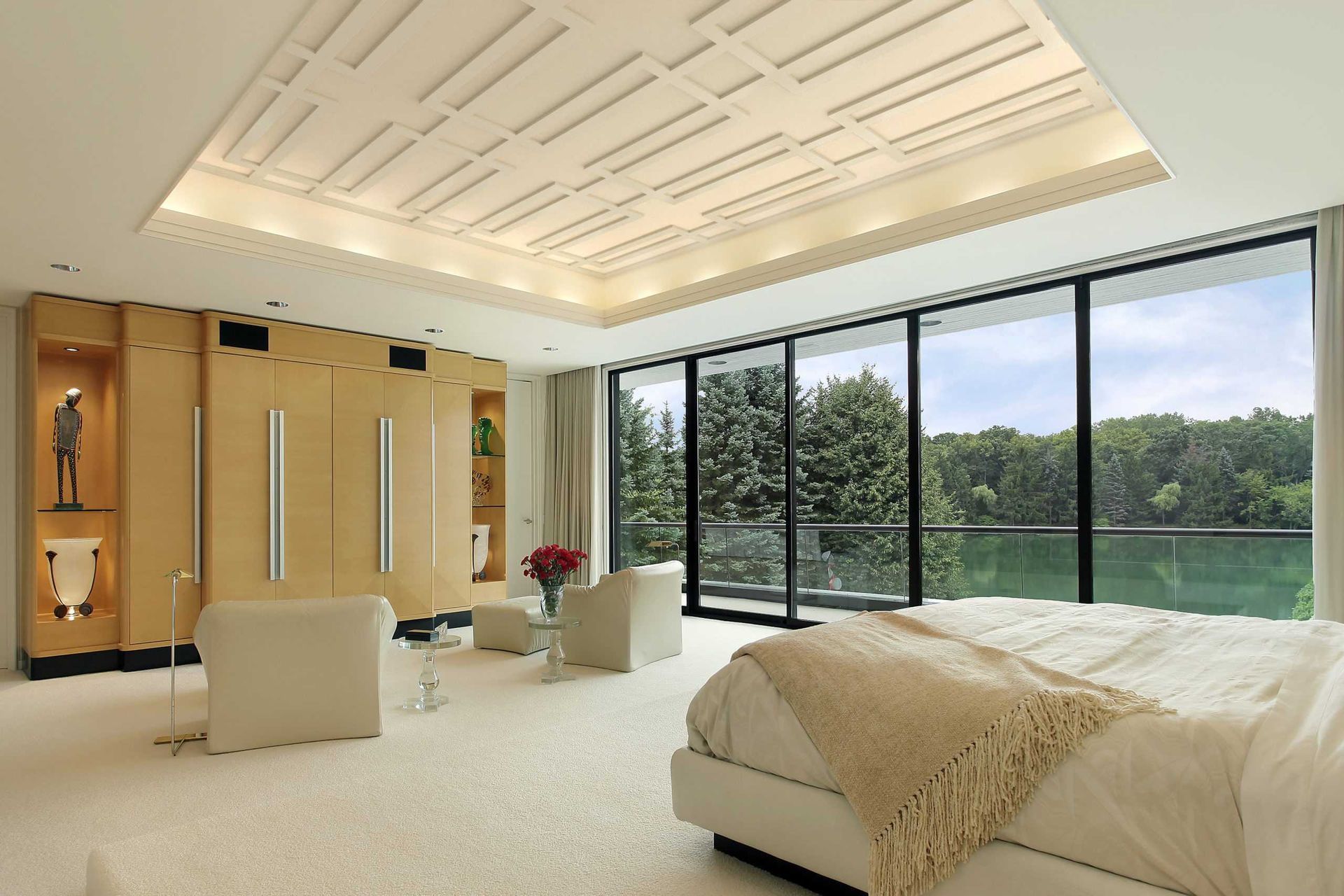 A modern bedroom with light wood walls, cream carpet, a white bed, and large floor-to-ceiling windows overlooking a forest.