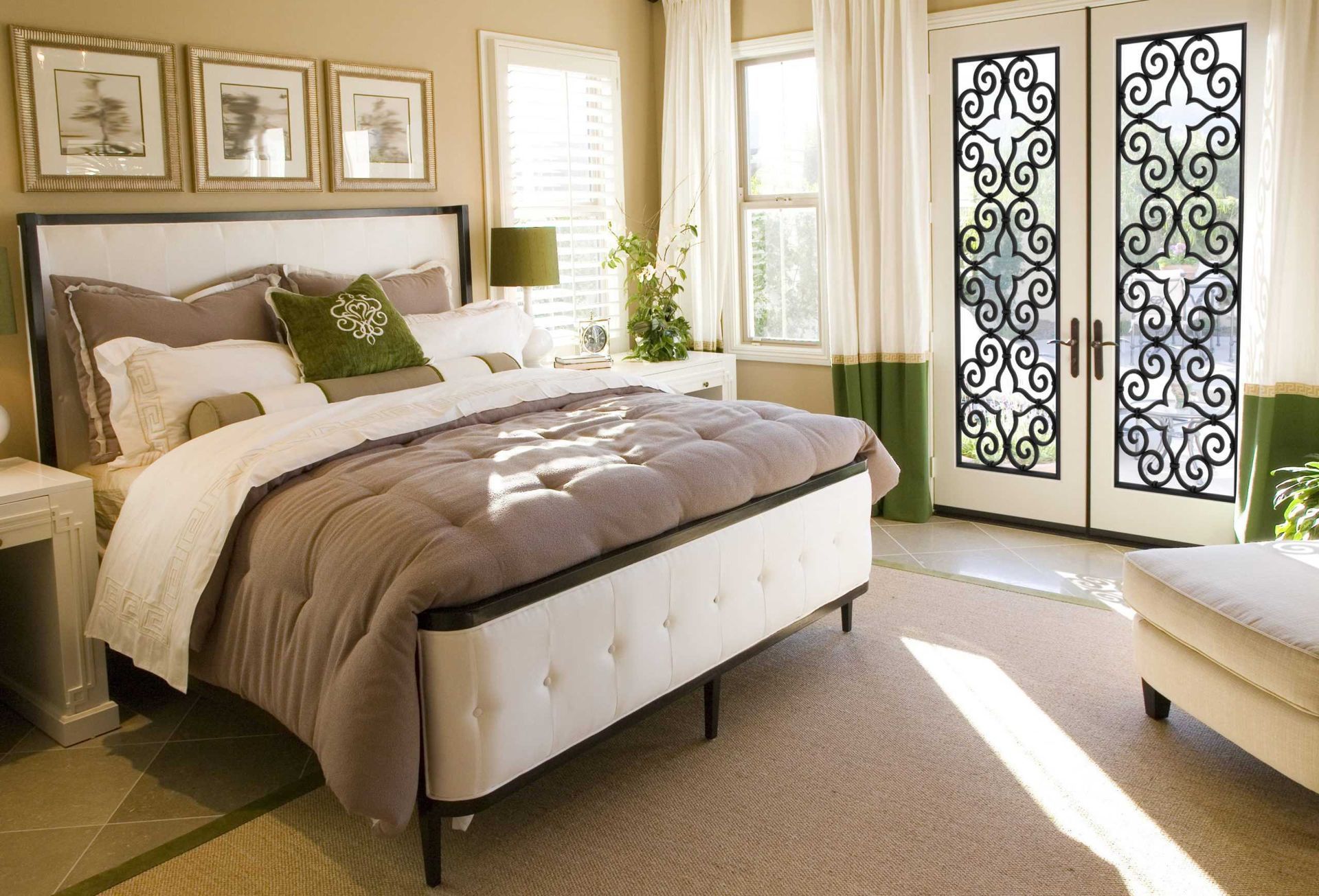 A bright bedroom featuring a tufted bed, white walls, and French doors with decorative iron grilles.