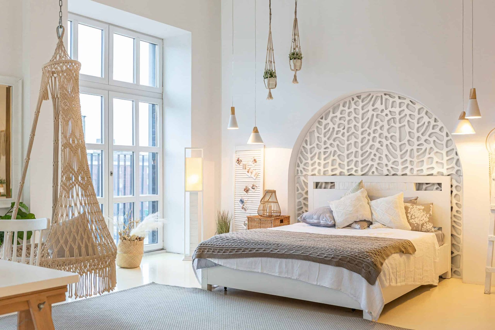 A bright bedroom with white walls, a bed against a circular patterned headboard, and a hanging macramé chair by the window.