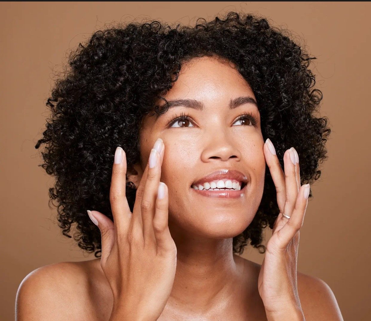A woman with curly hair is smiling and touching her face