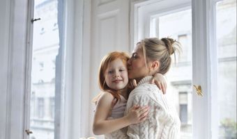 A woman is hugging a little girl in front of a window.