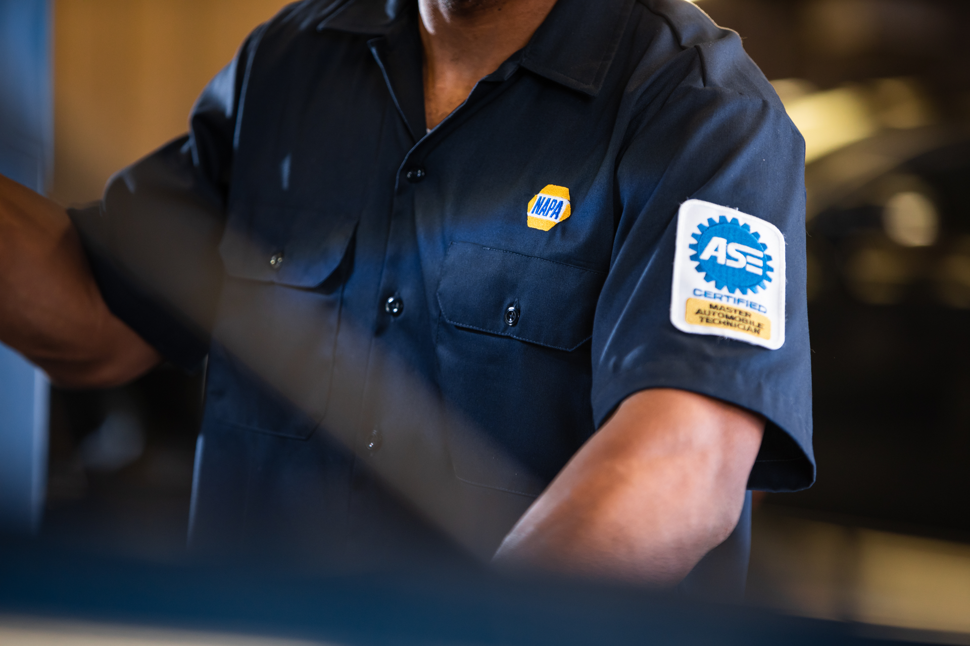Mechanic in a blue shirt with an ASE certification patch on sleeve, working in a shop.