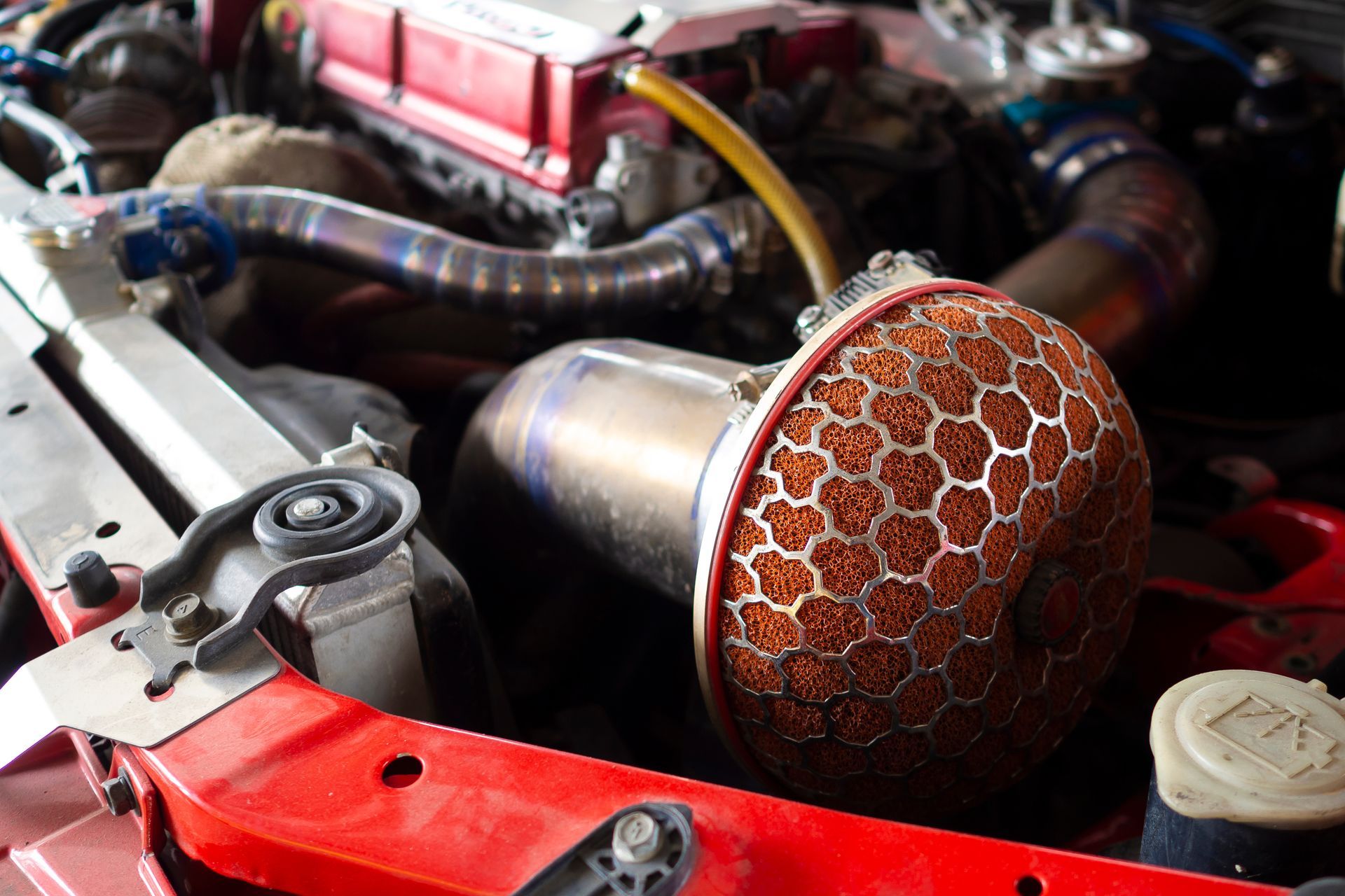 Close-up of a car engine bay, showing a red air filter, stainless steel tubing, and red engine components.