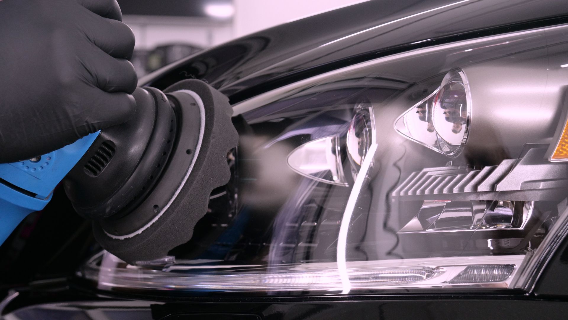 Black-gloved hand using an electric polisher on a car headlight to restore clarity.