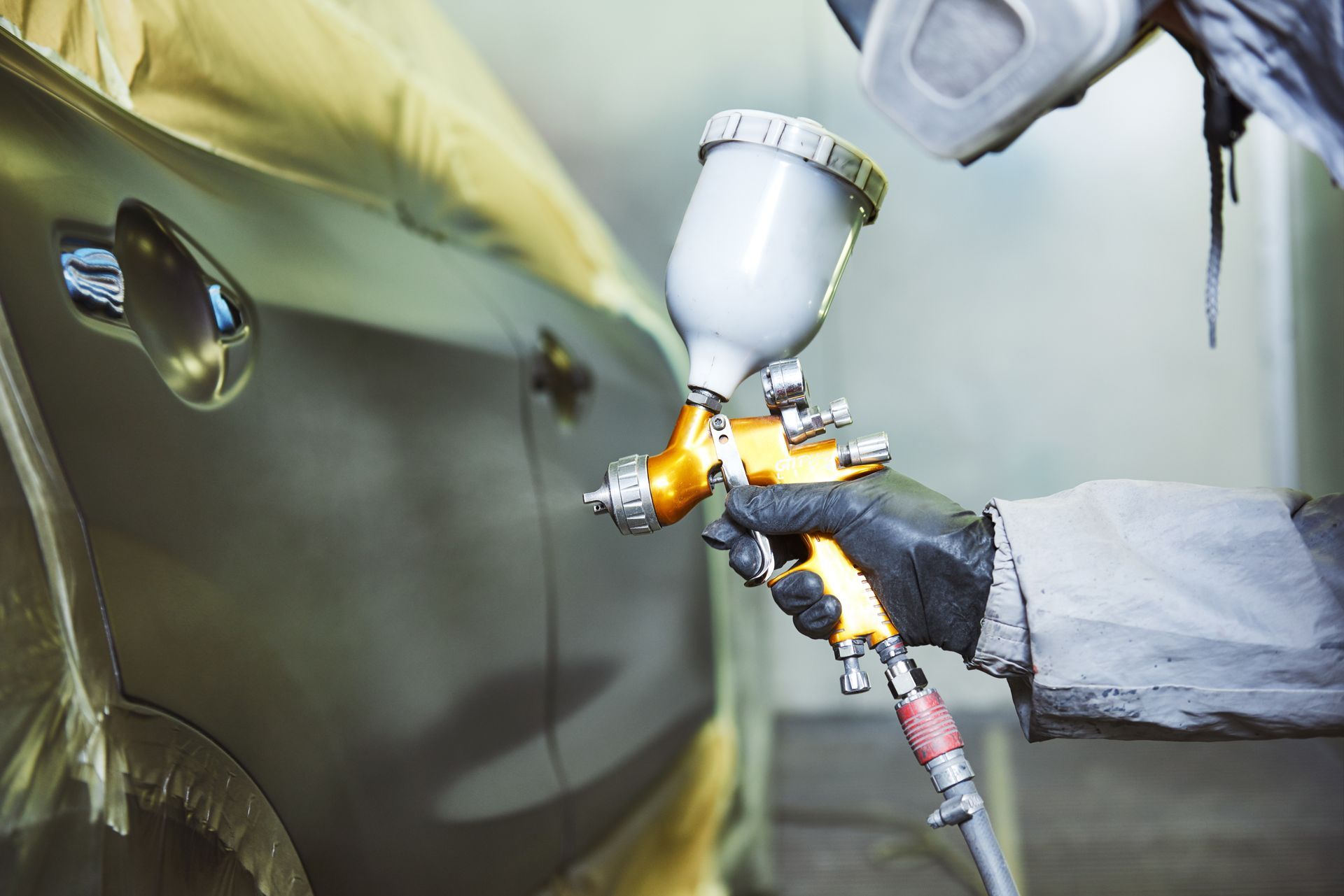 Person in protective gear sprays paint on a car with a spray gun in a body shop.