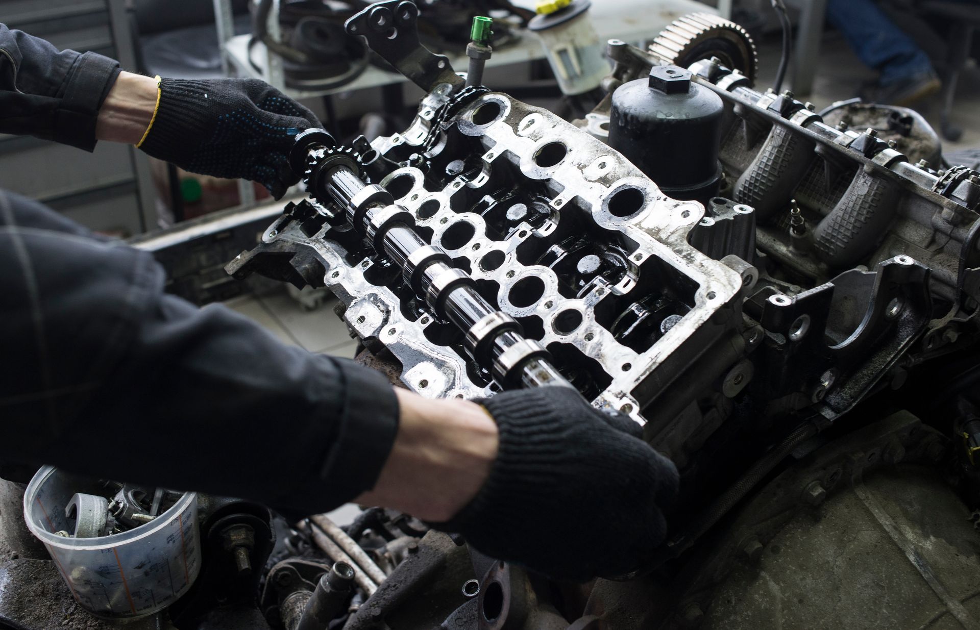 Hands wearing black gloves working on an engine cylinder head in a repair shop.