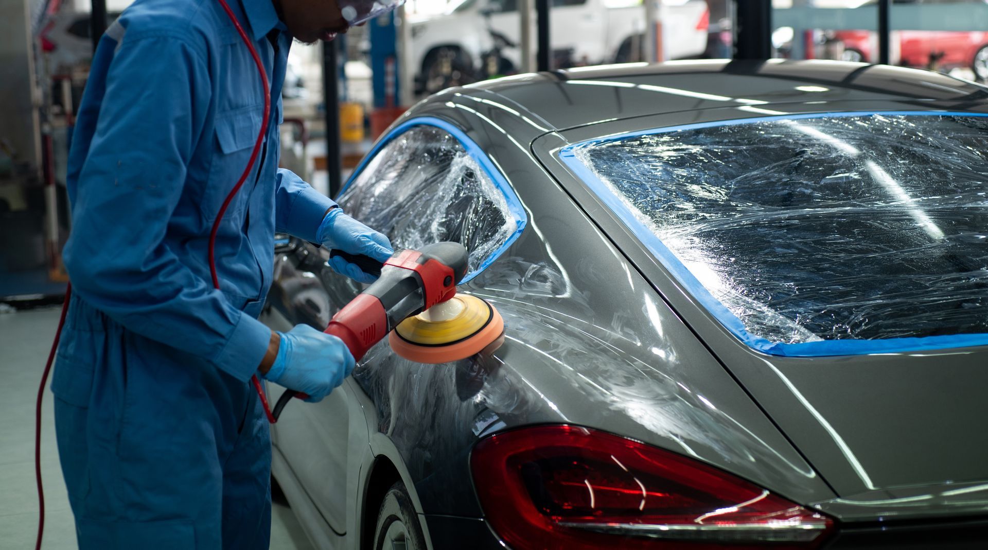 Person in blue coveralls polishes a gray car with an electric buffer in a garage.