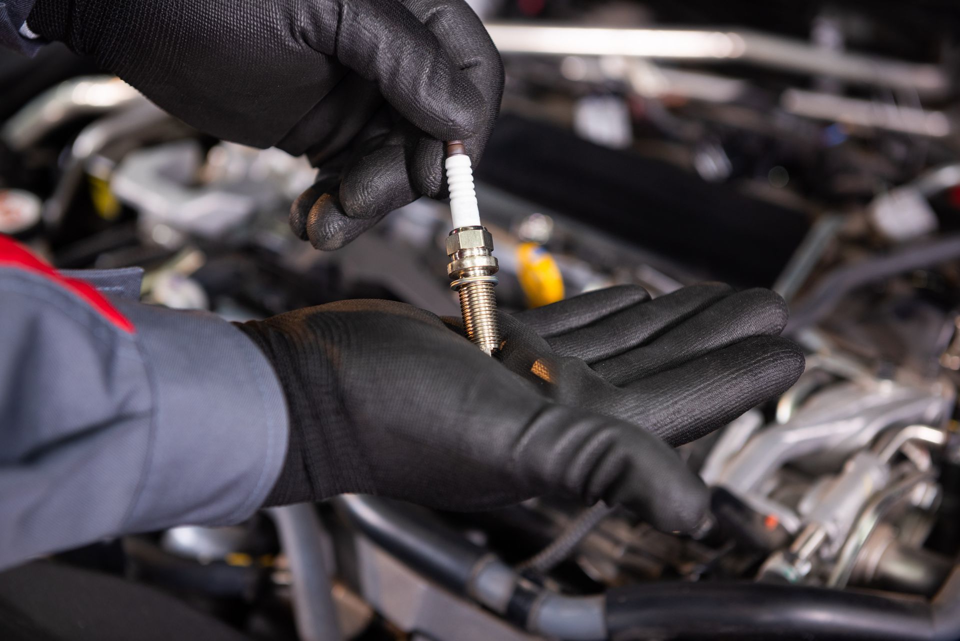 A mechanic in black gloves holding a spark plug in front of a car engine.
